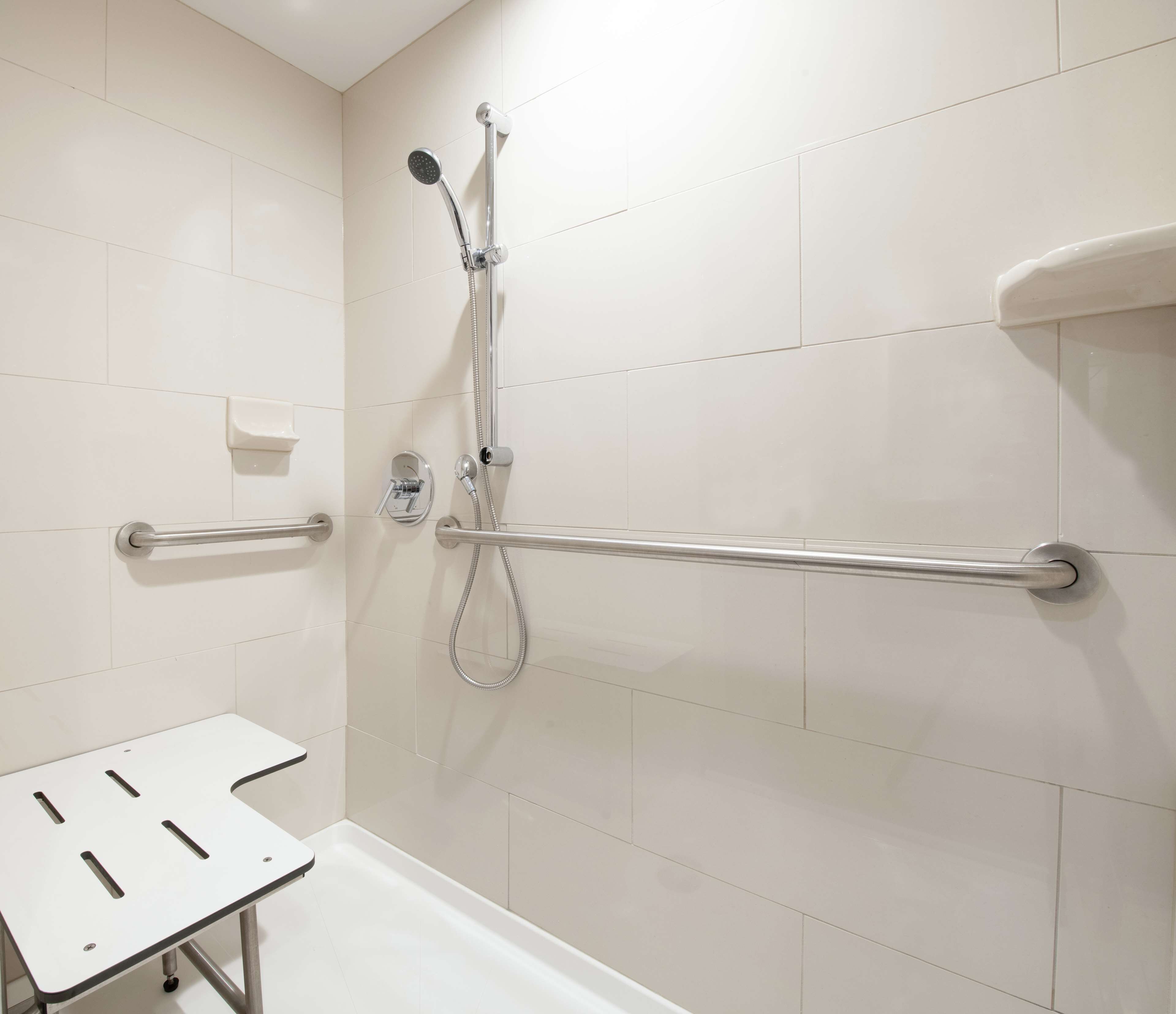 A modern bathroom with white tiles, a shower with a handheld showerhead, a grab bar, and a white bathtub.