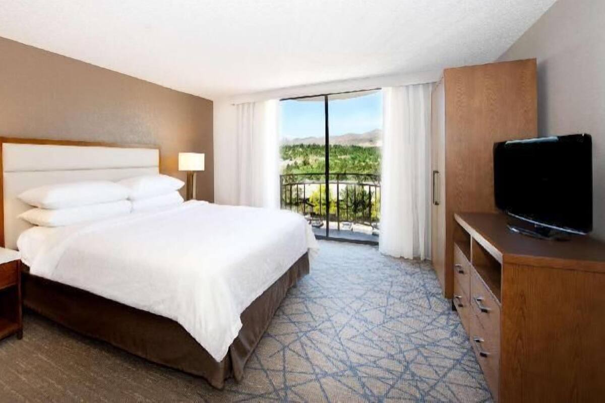 A hotel room with a bed, a TV, a nightstand, a dresser, and a balcony with a view.