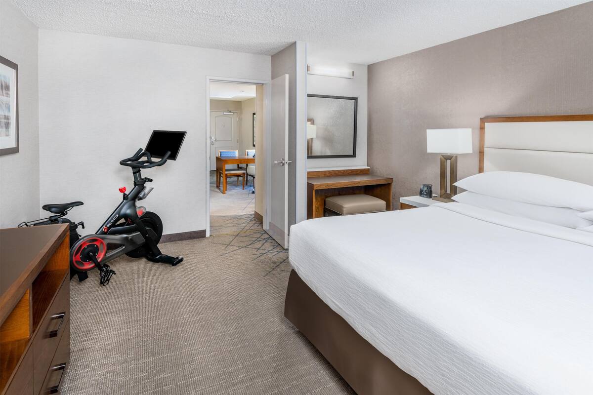 A hotel room with a bed, a desk, a chair, an exercise bike, and a door leading to a bathroom.