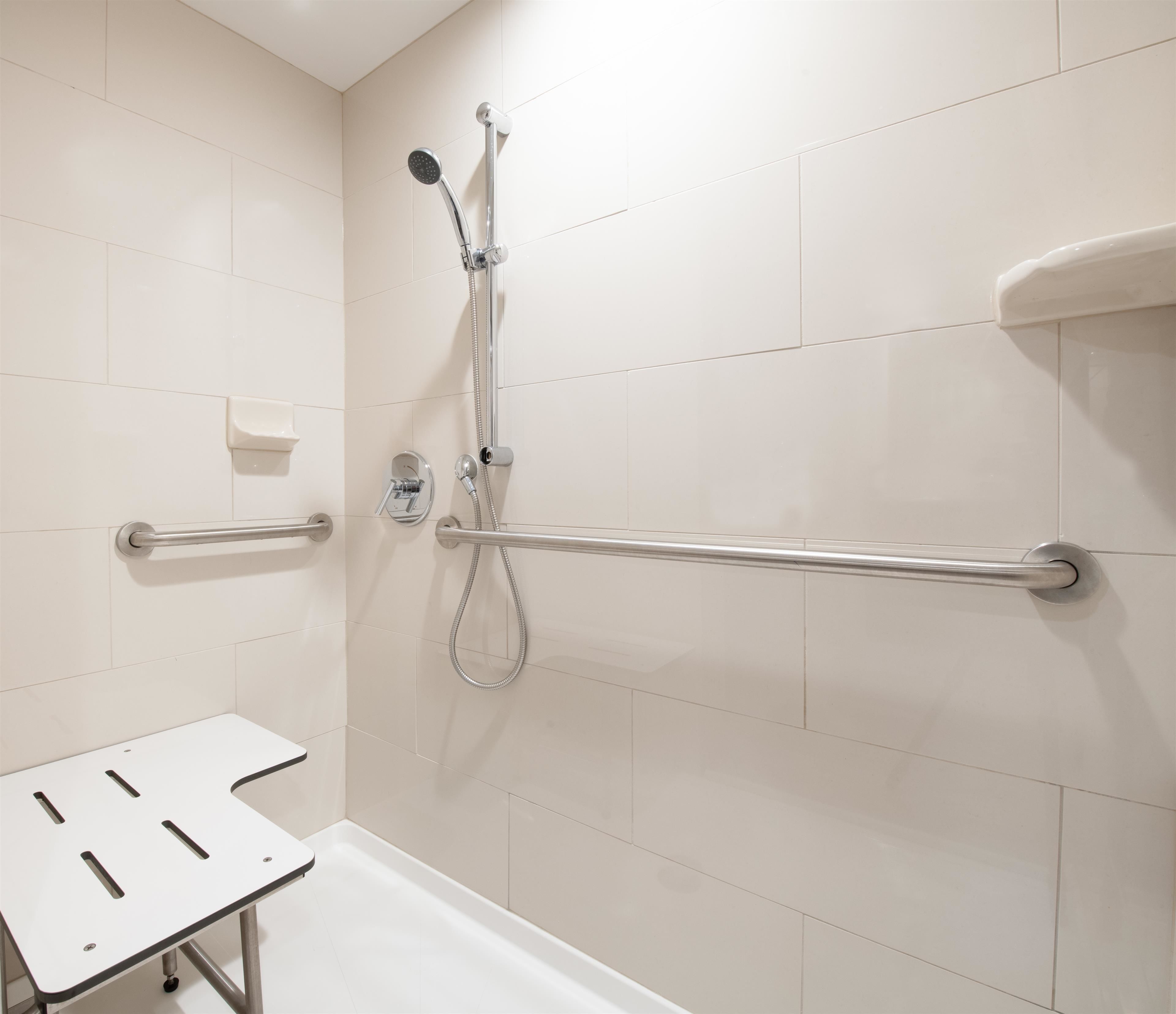 A bathroom with white tiles, a shower with a handheld showerhead, a grab bar, and a white sink.