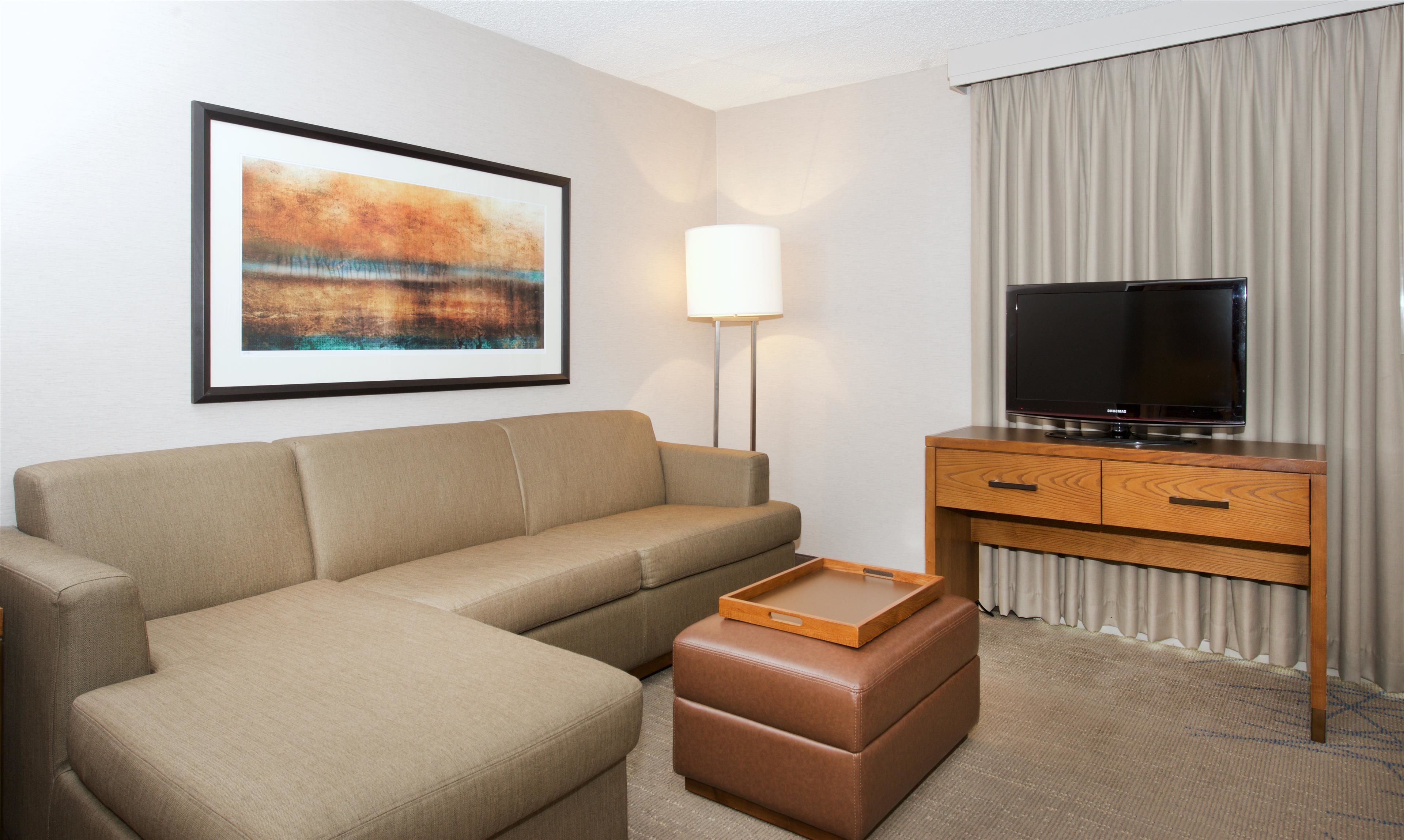 A hotel room with a beige sofa, a wooden TV stand, a flat-screen TV, a framed abstract painting, and a floor lamp.