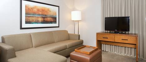 A hotel room with a beige sofa, a wooden TV stand, a flat-screen TV, a framed abstract painting, and a floor lamp.