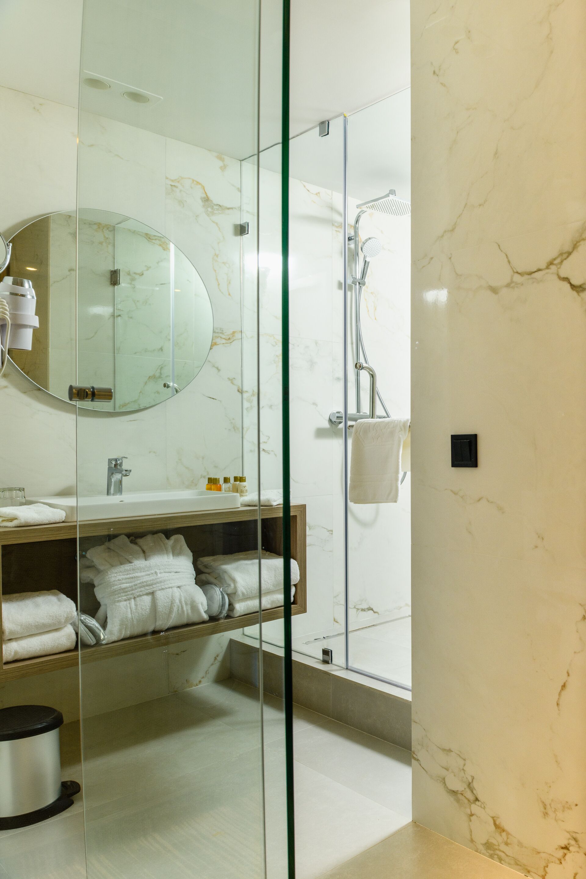 Un baño moderno con ducha envuelta en cristal, tocador de mármol y espejo redondo.