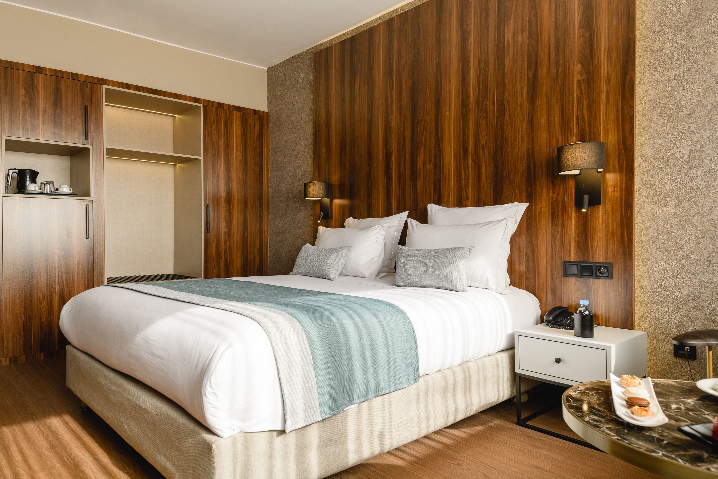A modern hotel room with a large bed, wooden panel walls, a nightstand with a lamp, and a small round table with snacks.