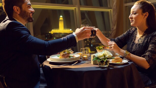 Una pareja brindando en una mesa con una vela, una copa de vino y un plato de postre.
