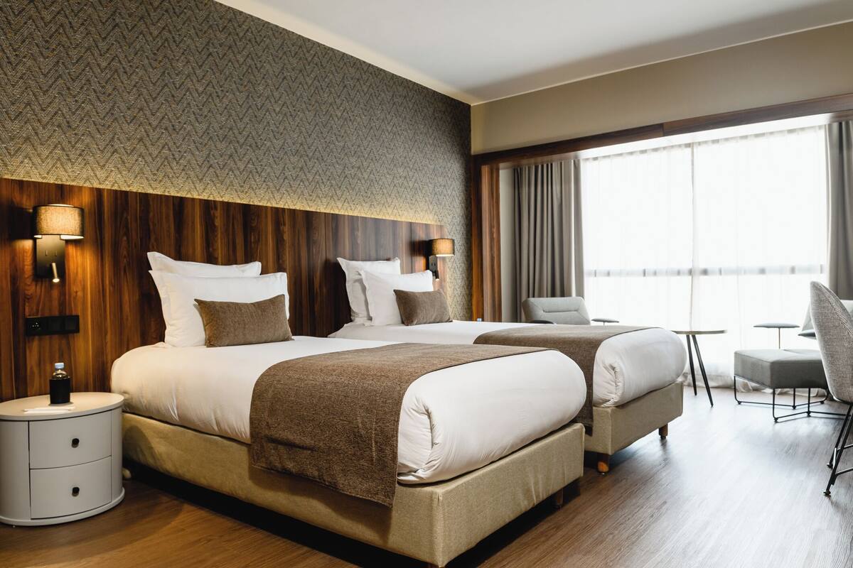 A hotel room with two beds, a wooden headboard, a nightstand, a lamp, a chair, and a large window with curtains.