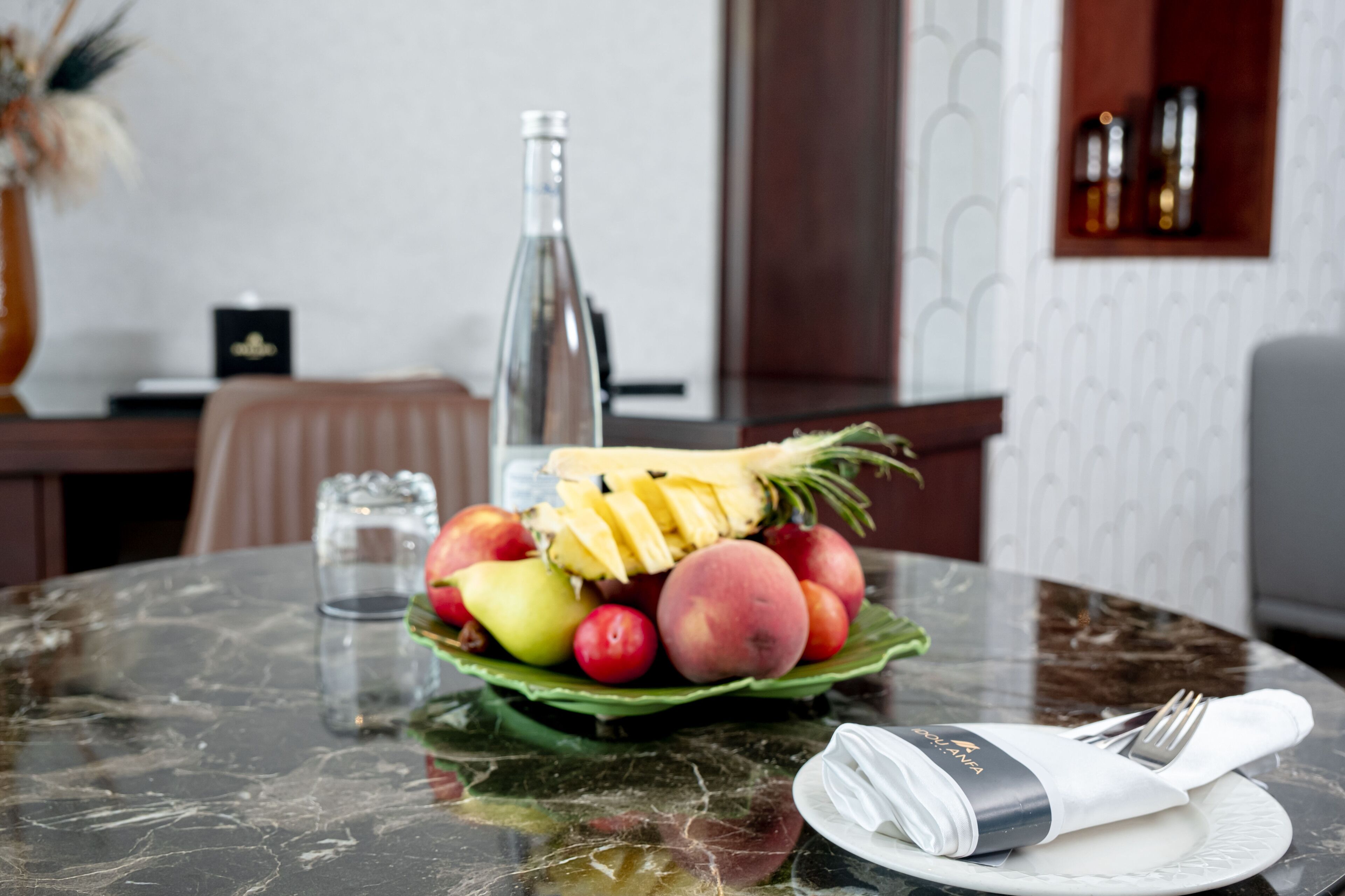 Una mesa de comedor con un frutero que contiene varias frutas, una botella y un mantel doblado con un tenedor.