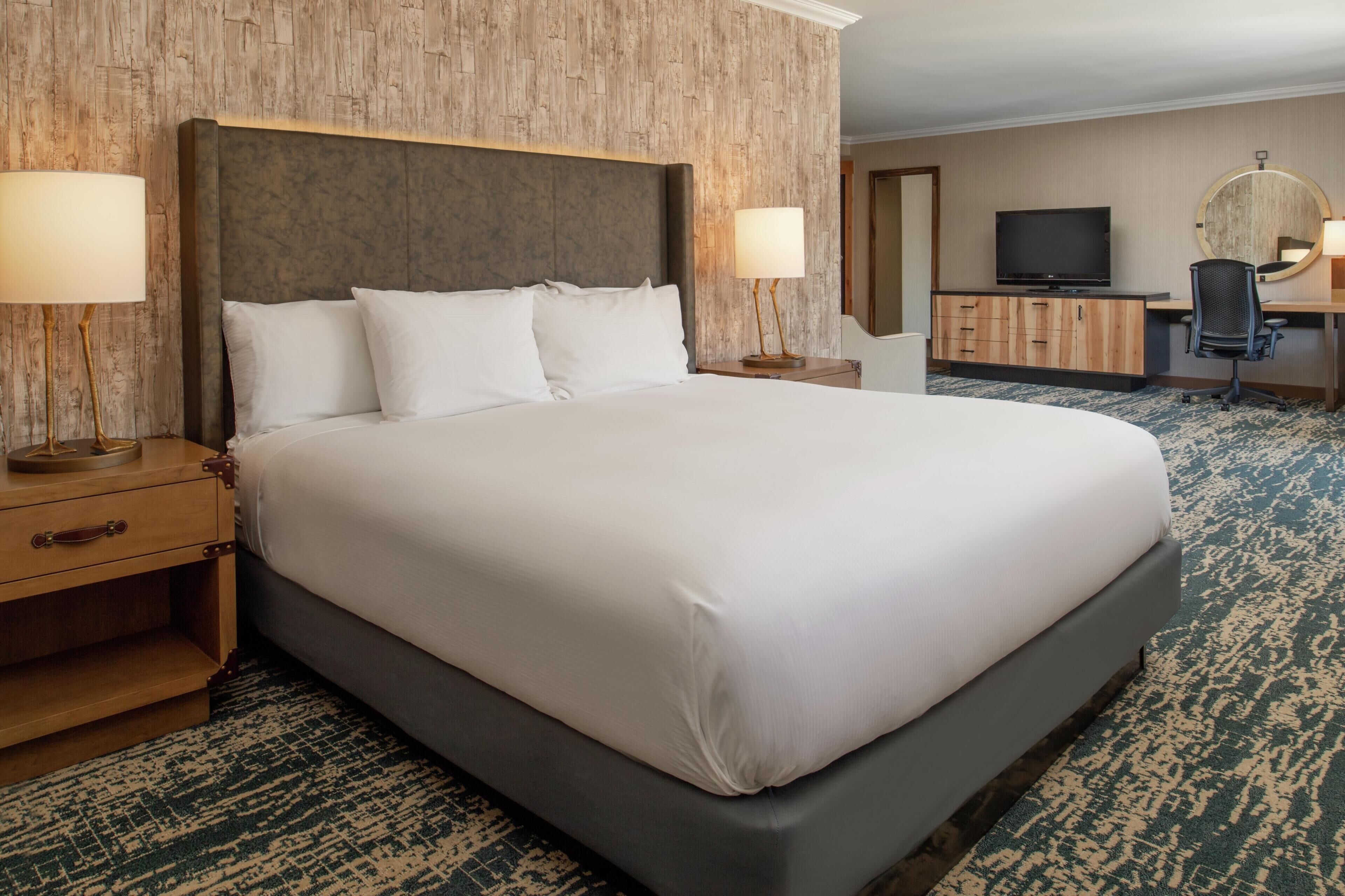 A hotel room with a large bed, two bedside tables with lamps, a desk with a chair, a television, and a mirror.