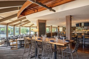 A spacious restaurant with a long wooden table, metal chairs, and a bar area with shelves of bottles.