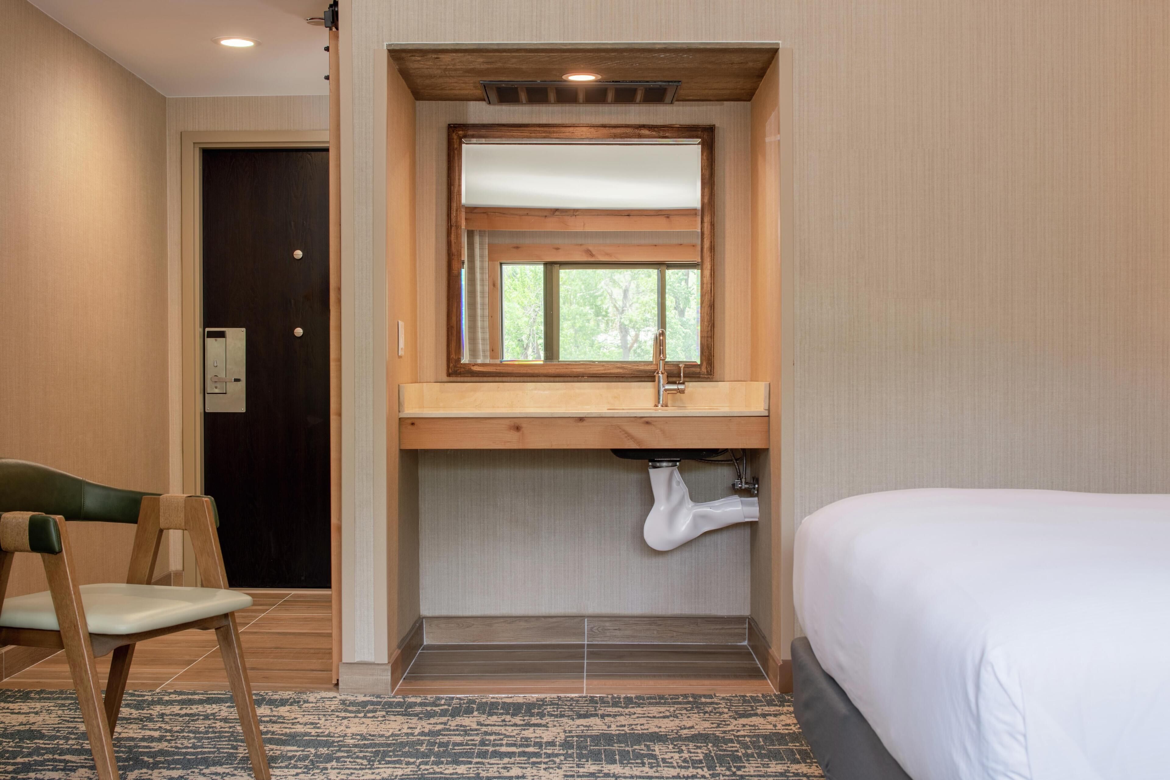 A hotel room with a bed, a wooden desk with a sink, a chair, and a window.