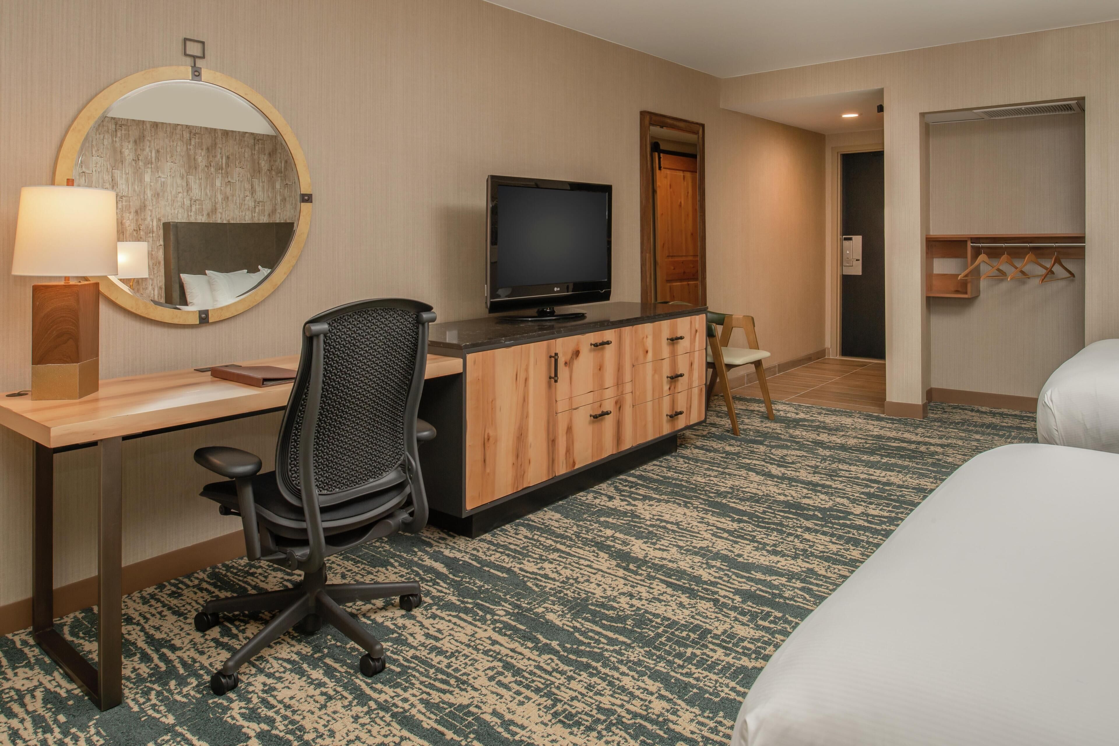 A hotel room with a bed, a desk with a computer, a television, a mirror, and a wardrobe.