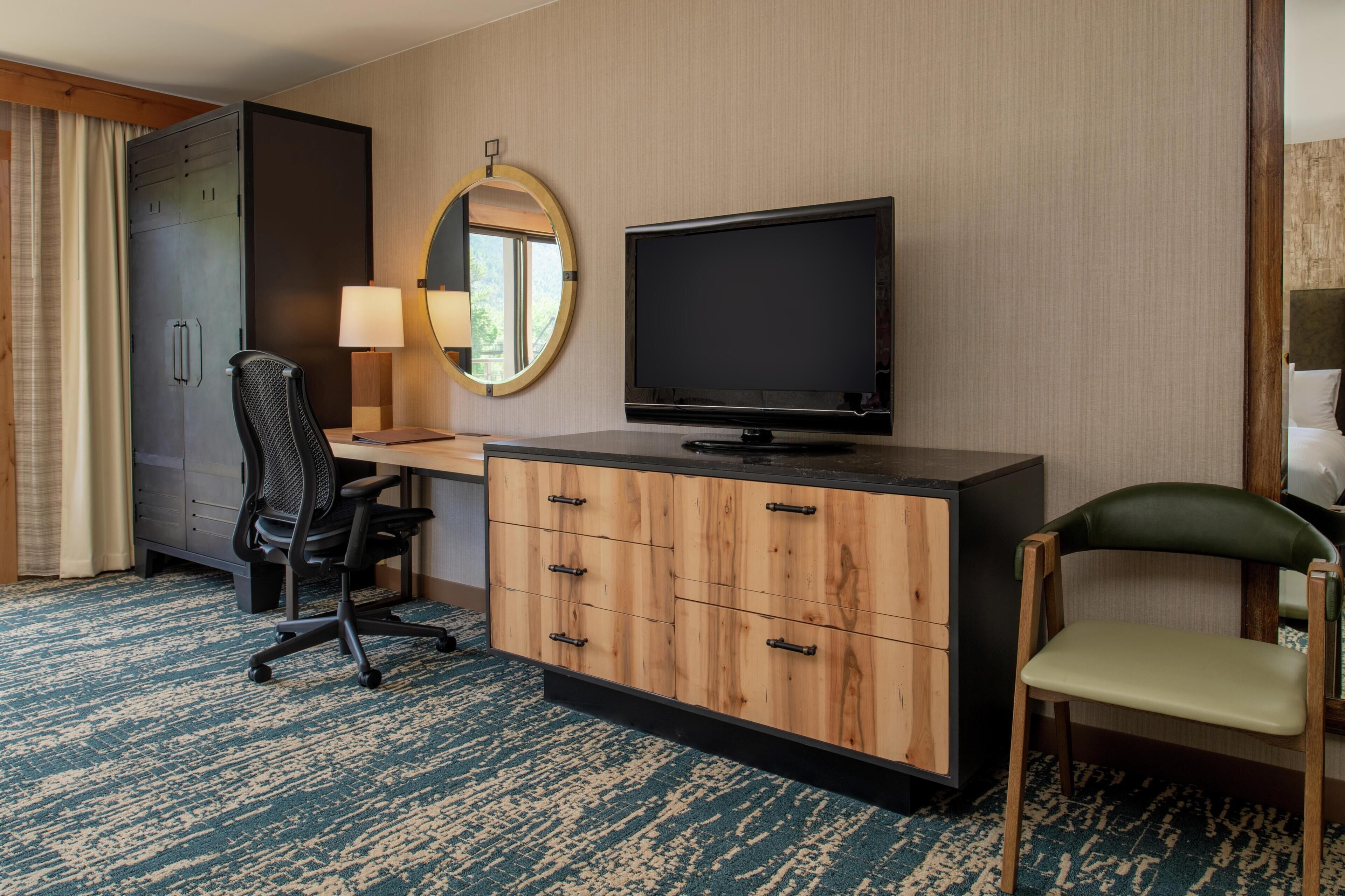 A hotel room with a desk, chair, television, wardrobe, and a mirror.