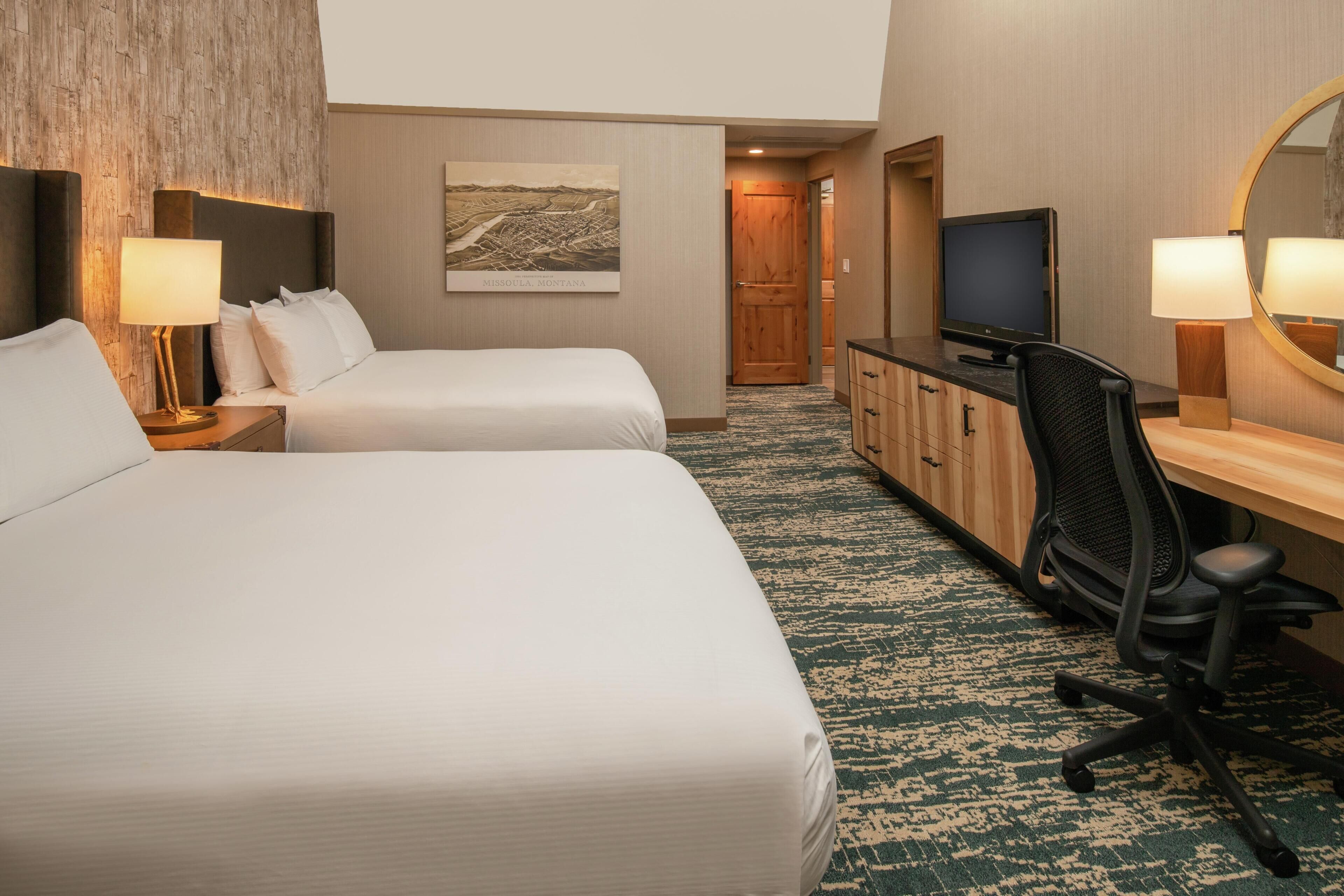 Ein Hotelzimmer mit zwei Betten, einem Schreibtisch mit Computer, einem Fernseher und einem Gemälde an der Wand.