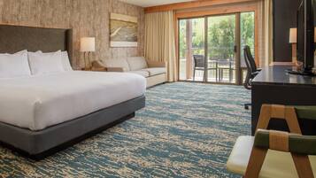 A hotel room with a large bed, a sofa, a desk with a computer, and a view of the outdoors through a sliding glass door.