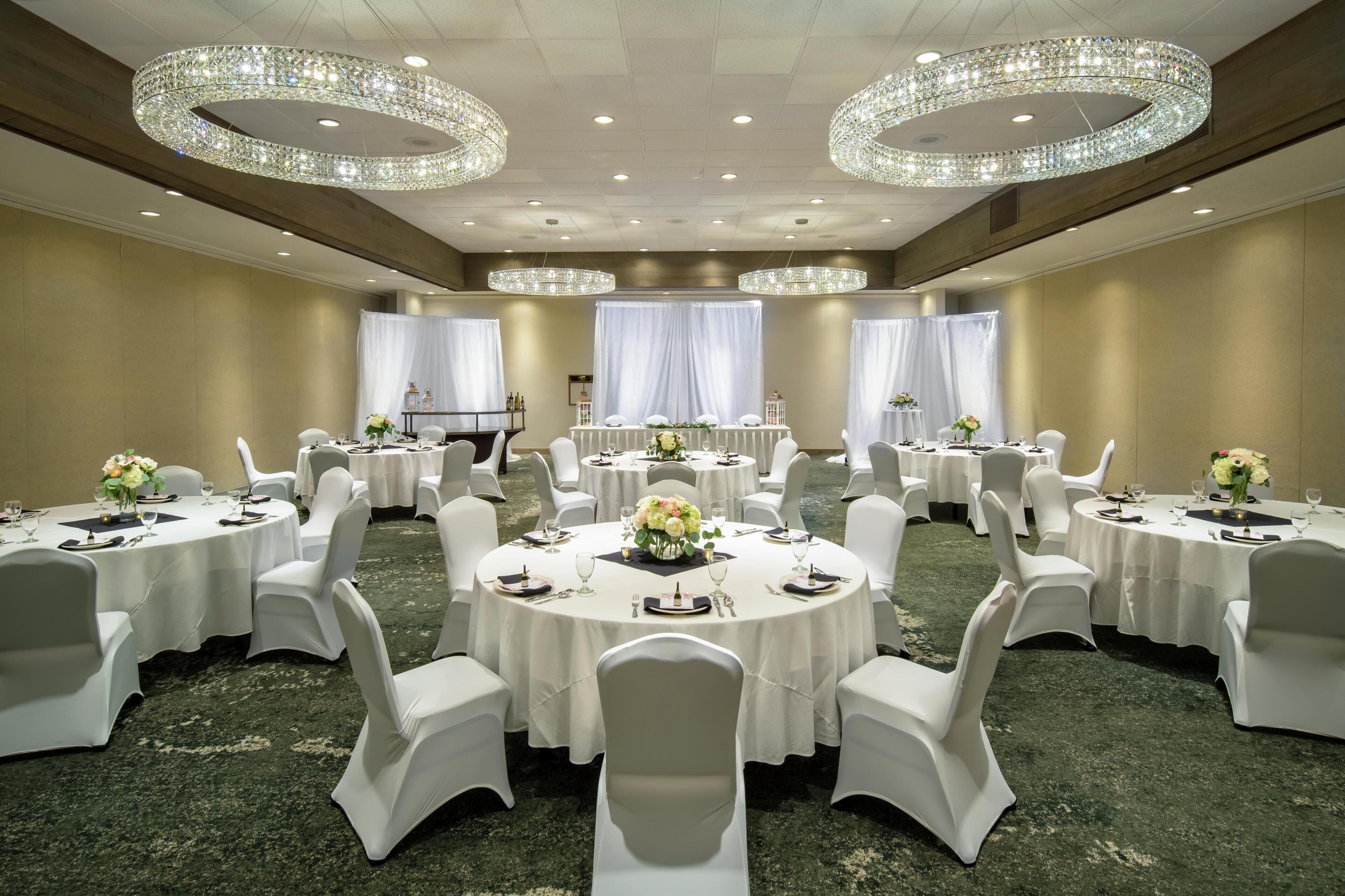 A banquet hall with round tables, white chairs, chandeliers, and a carpeted floor.