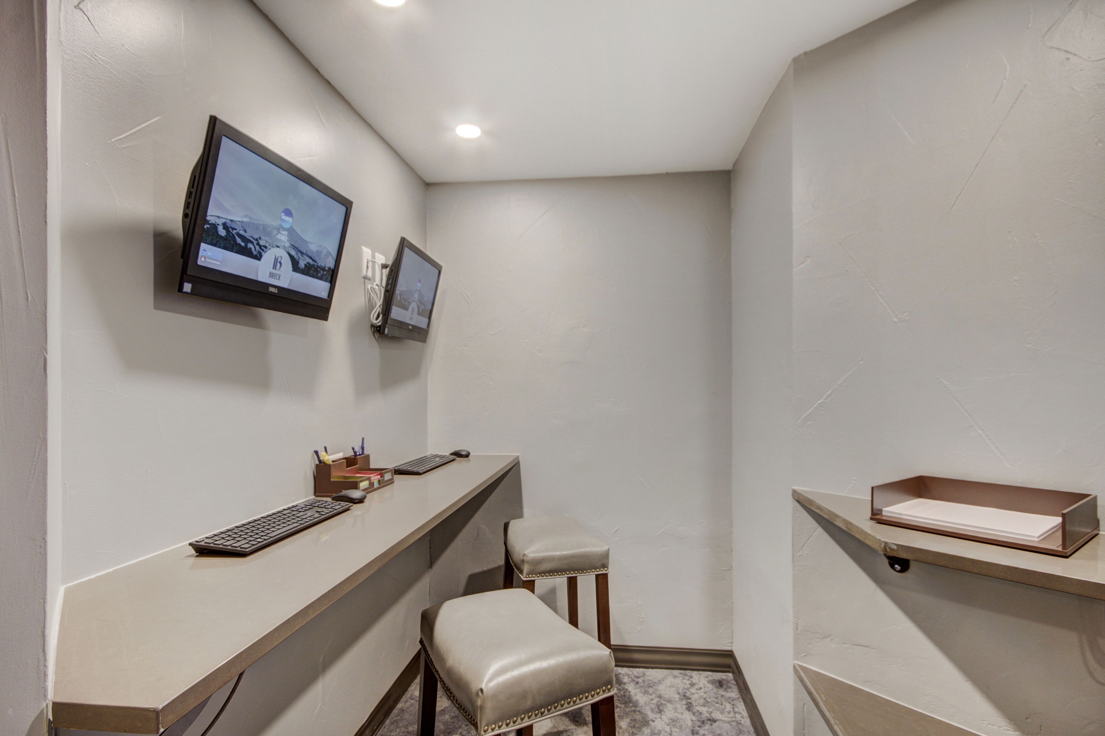 A small, modern office space with a desk, two chairs, a mounted television, and a shelf with a box.