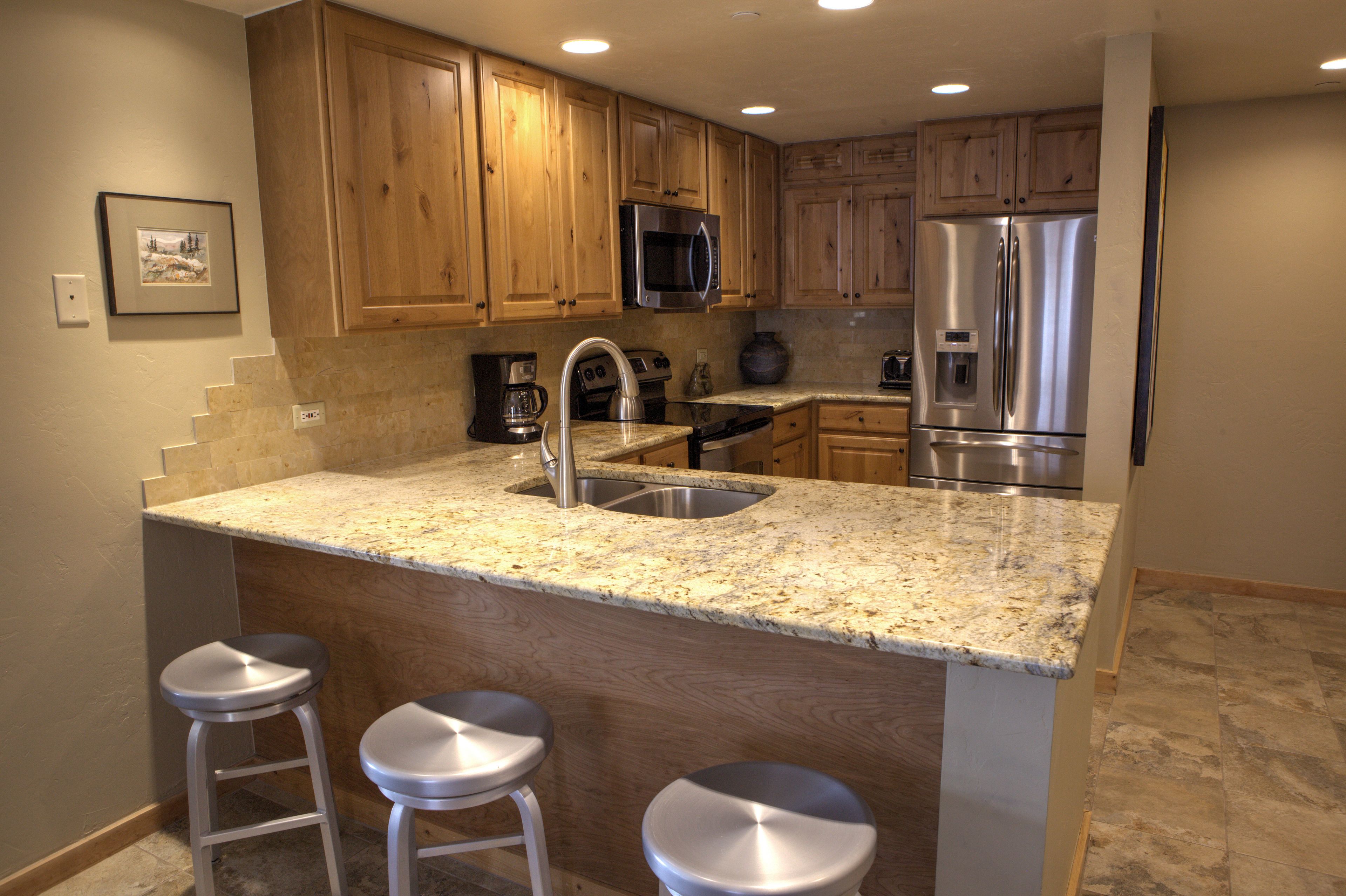 A kitchen with wooden cabinets, stainless steel appliances, a granite countertop, and three bar stools.