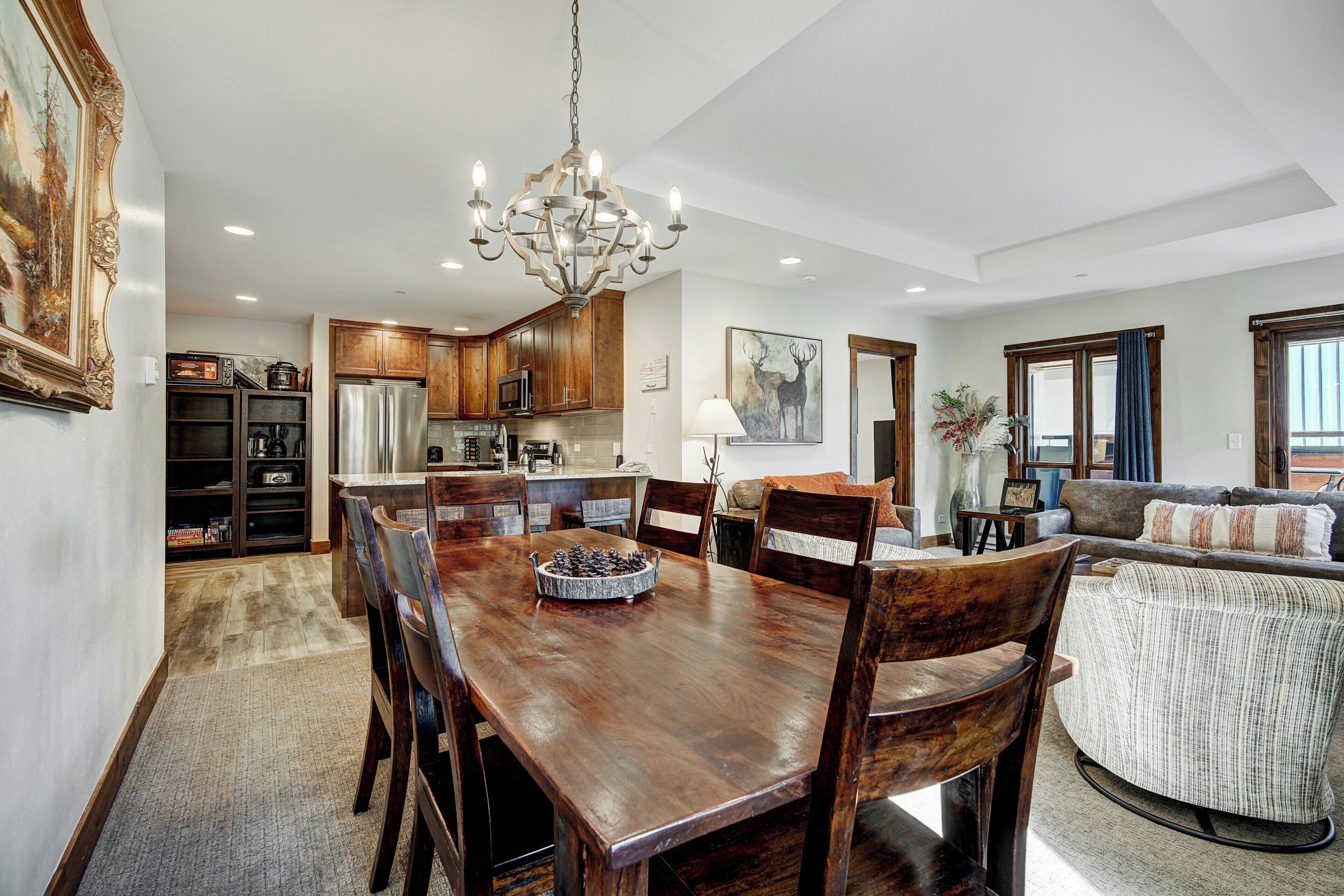 A spacious dining area with a wooden table and chairs, a chandelier, and a kitchen in the background.