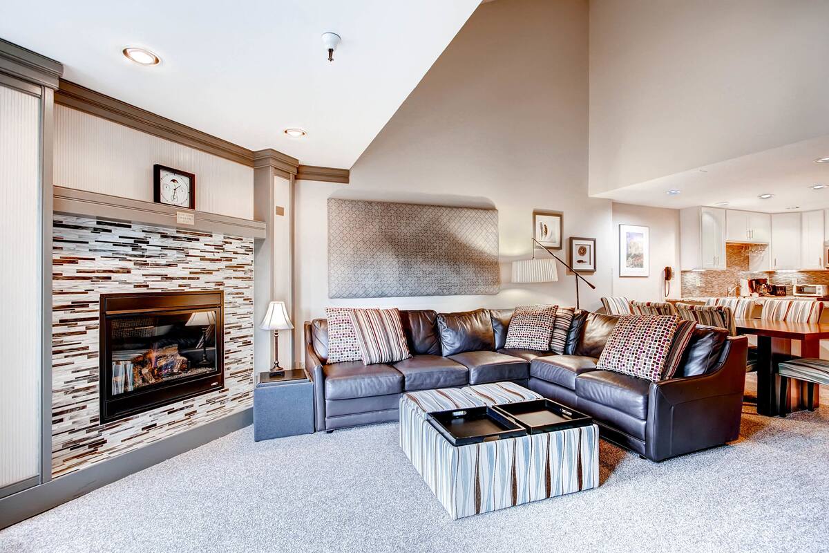 A modern living room with a sectional sofa, a fireplace, and a dining area in the background.