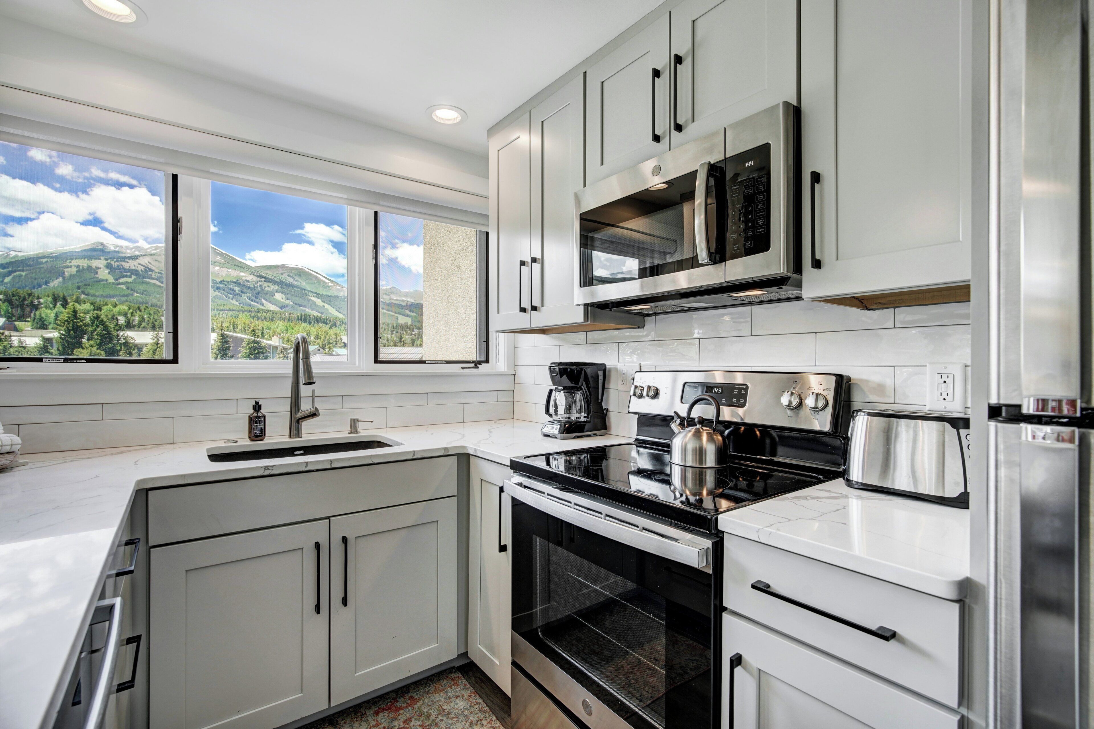 Una cocina moderna con gabinetes blancos, electrodomésticos de acero inoxidable, un amplio ventanal con vistas a la montaña y un horno empotrado.