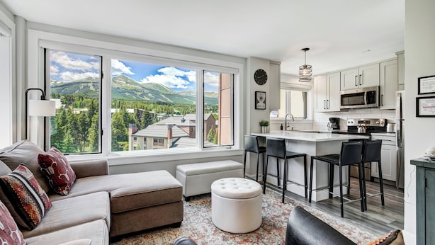 A modern living room with a large window, a sofa, ottoman, and a kitchen area with bar stools. - Village at Breckenridge, A Vail Resorts Property (Breckenridge)