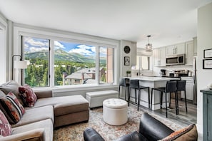 A modern living room with a large window, a sofa, ottoman, and a kitchen area with bar stools.