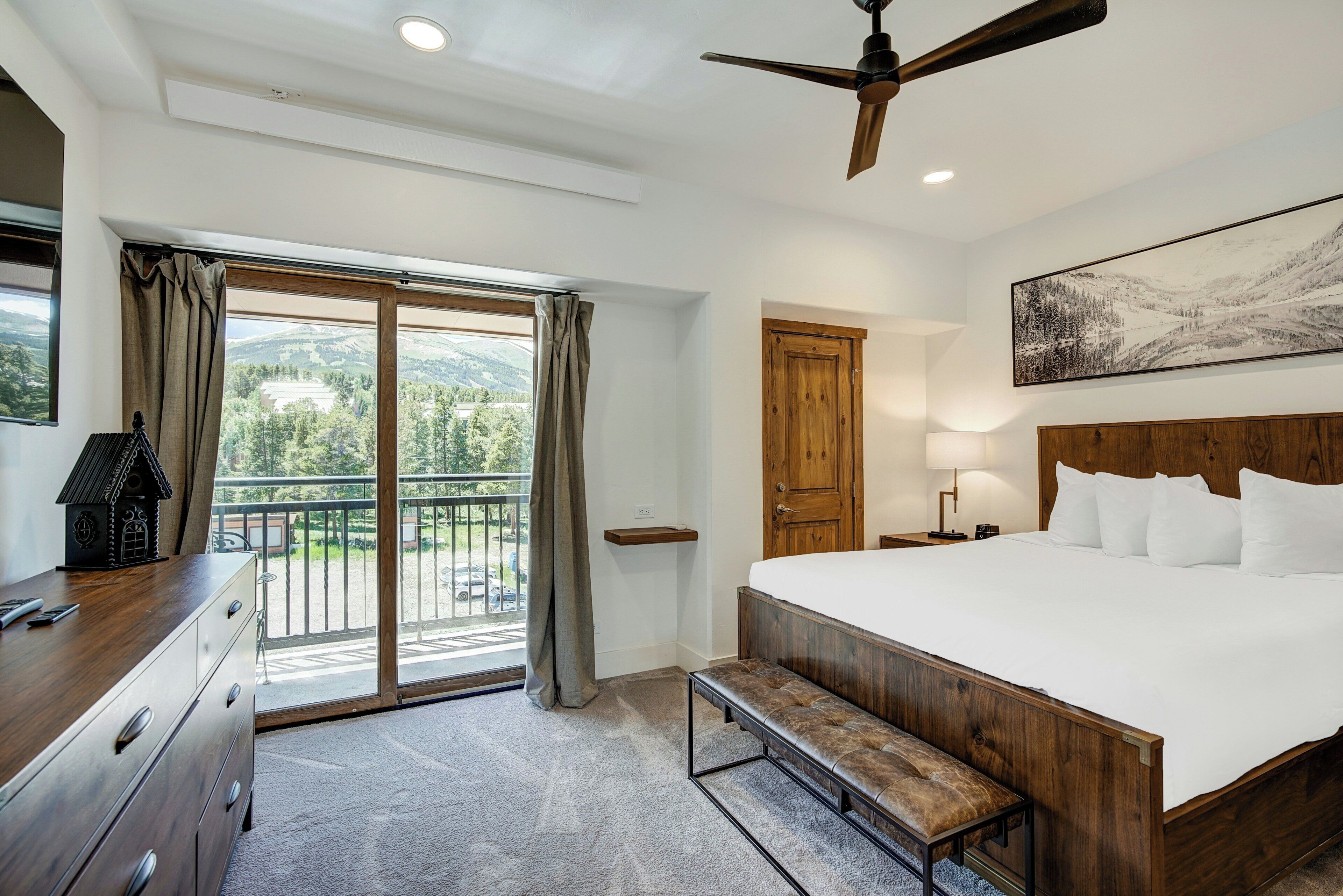 A bedroom with a large bed, wooden headboard, a bench, a ceiling fan, a wooden dresser, a desk with a lamp, a painting on the wall, and a sliding glass door leading to a balcony.