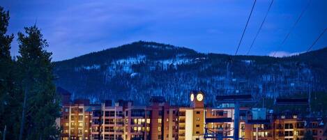 A multi-story hotel with a clock tower, ski lift, and mountainous backdrop.