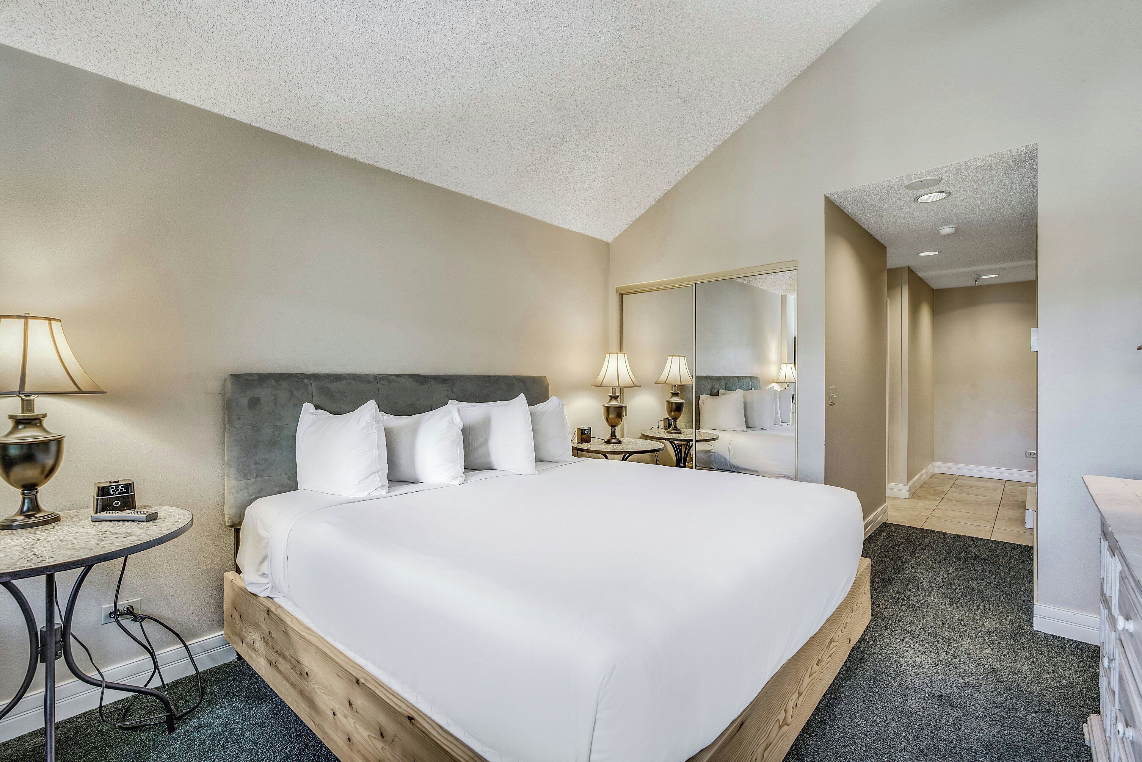 Una habitación de hotel con una cama grande, mesitas de noche, un espejo y una pequeña mesa con lámpara.