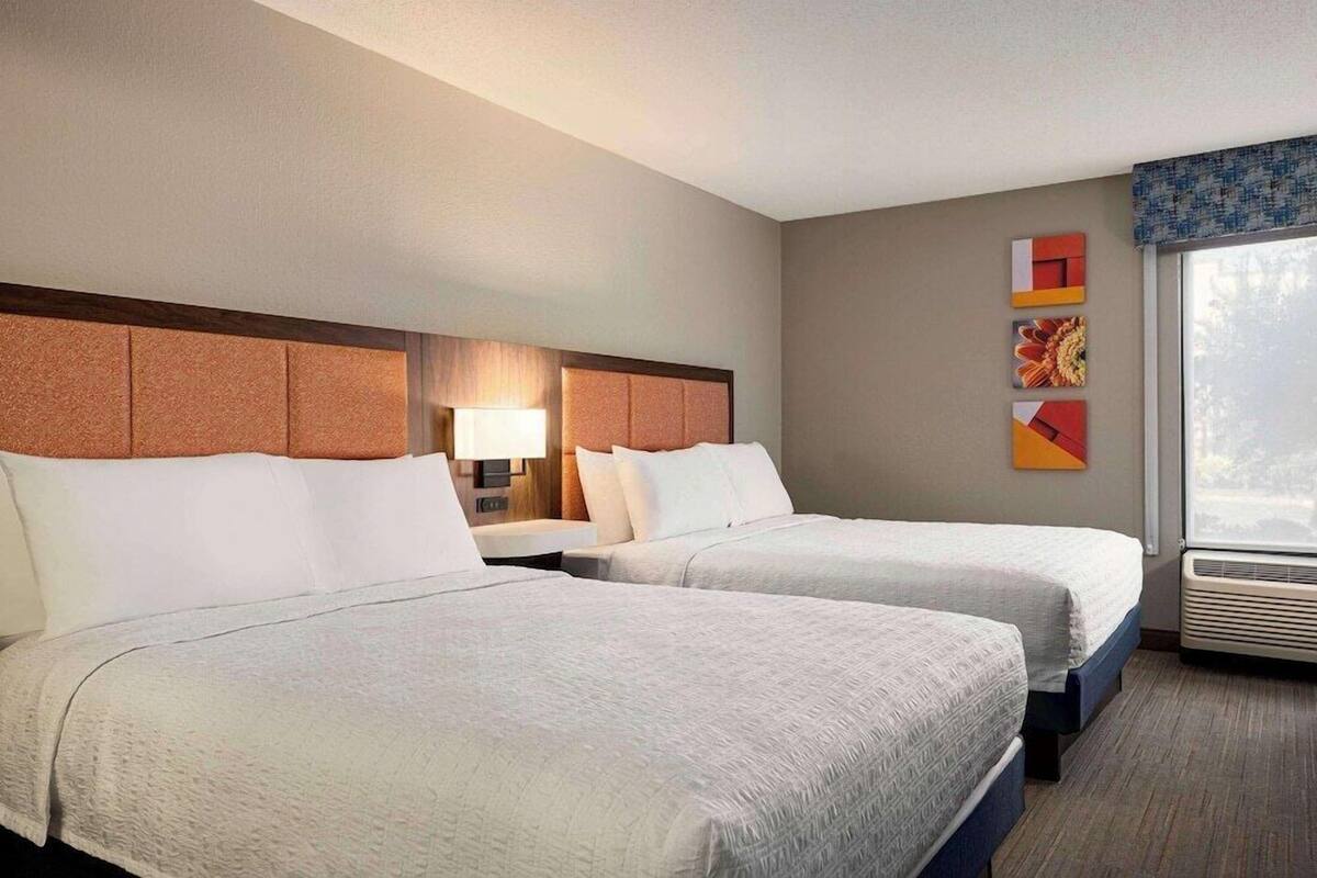 A hotel room with two beds, a nightstand, a lamp, a window with a view, and wall art.