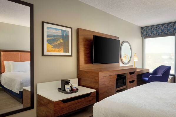 Habitación de hotel con cama, escritorio con cafetera, televisor de pantalla plana, espejo y ventana con vistas.