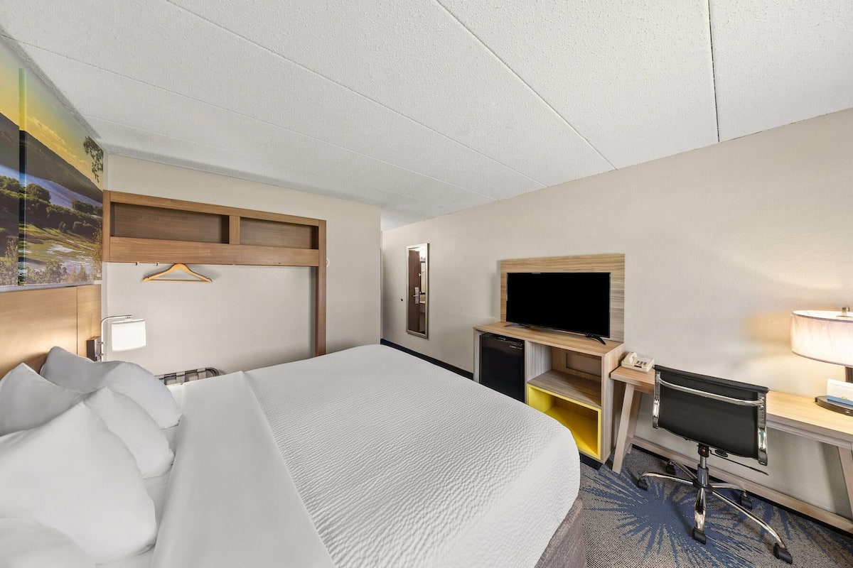 Ein Hotelzimmer mit einem großen Bett, einem Schreibtisch mit Computer, einem Fernseher und einem an der Wand befestigten Fernseher.