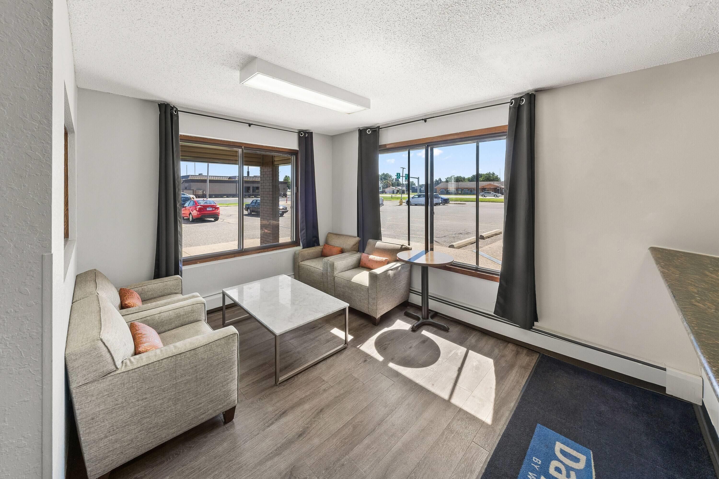 A modern hotel room with a sofa, armchair, and coffee table. Large windows offer a view of the parking lot and street.