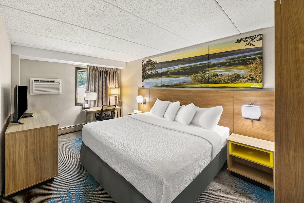 Ein Hotelzimmer mit einem großen Bett, einem Schreibtisch, einem Sessel, einem Fernseher, einer Klimaanlage und einem Landschaftsbild.