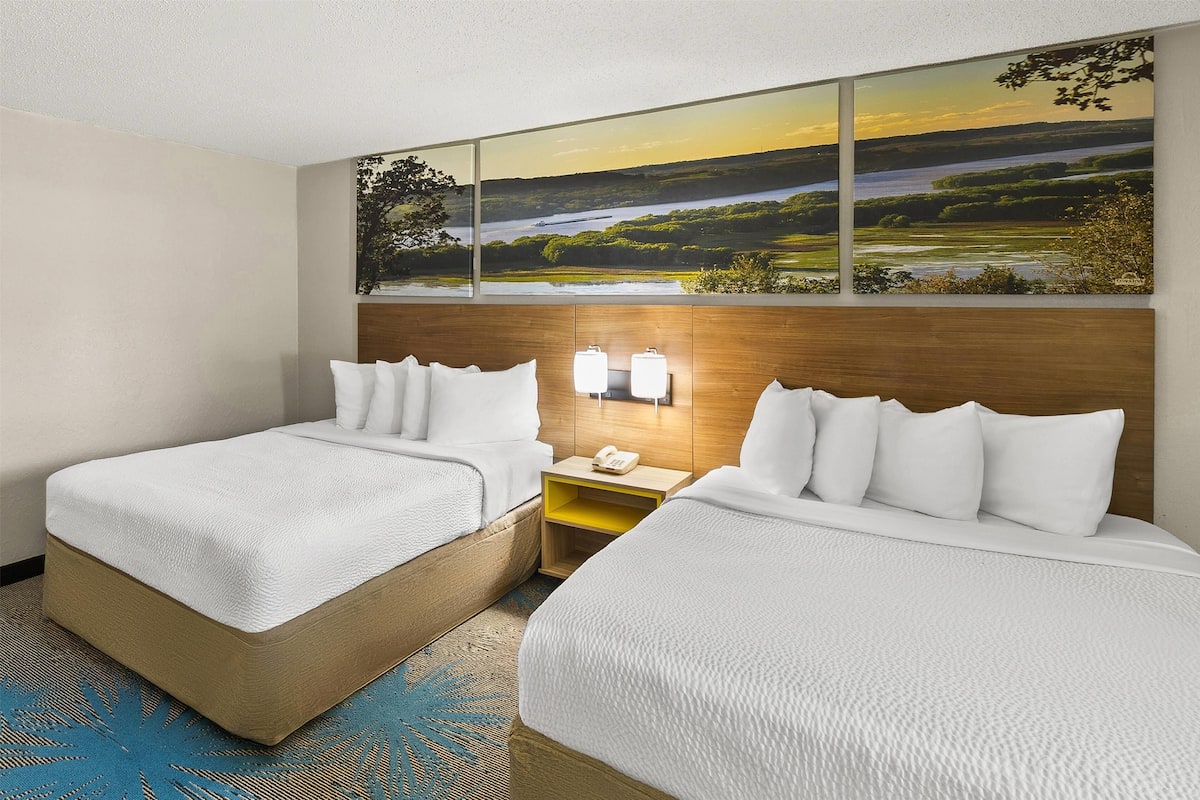 Ein Hotelzimmer mit zwei Betten, einem großen Landschaftsbild über dem Kopfteil, einem Nachttisch mit Telefon und einem blauen Teppich mit Muster.