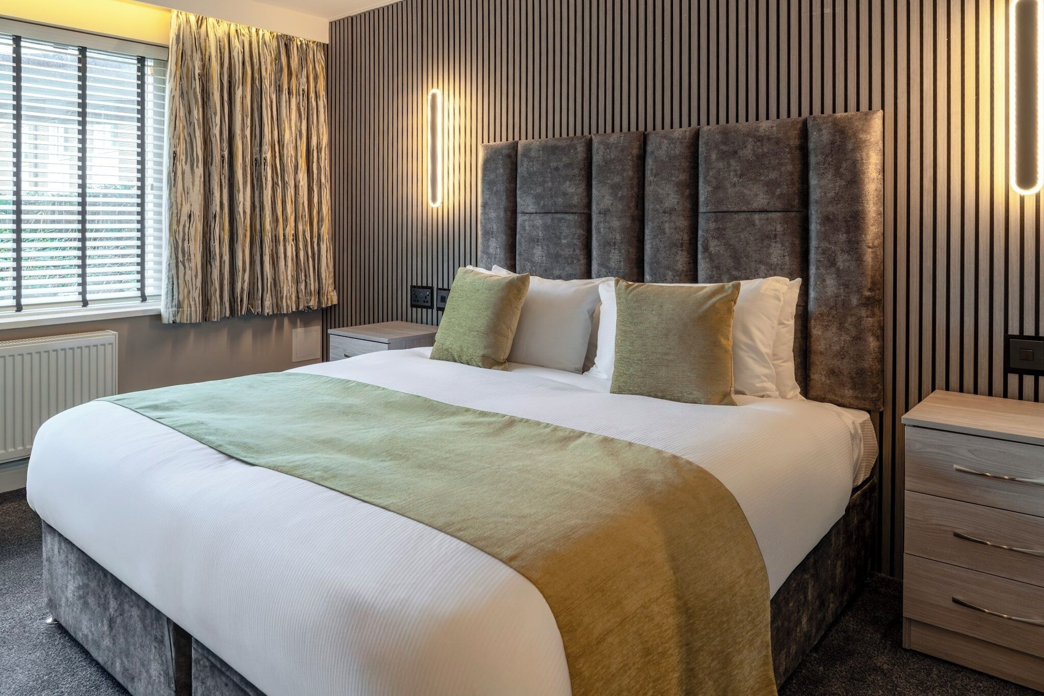 Quarto de hotel moderno com uma cama grande, uma mesa de cabeceira e uma luminária fixada na parede.