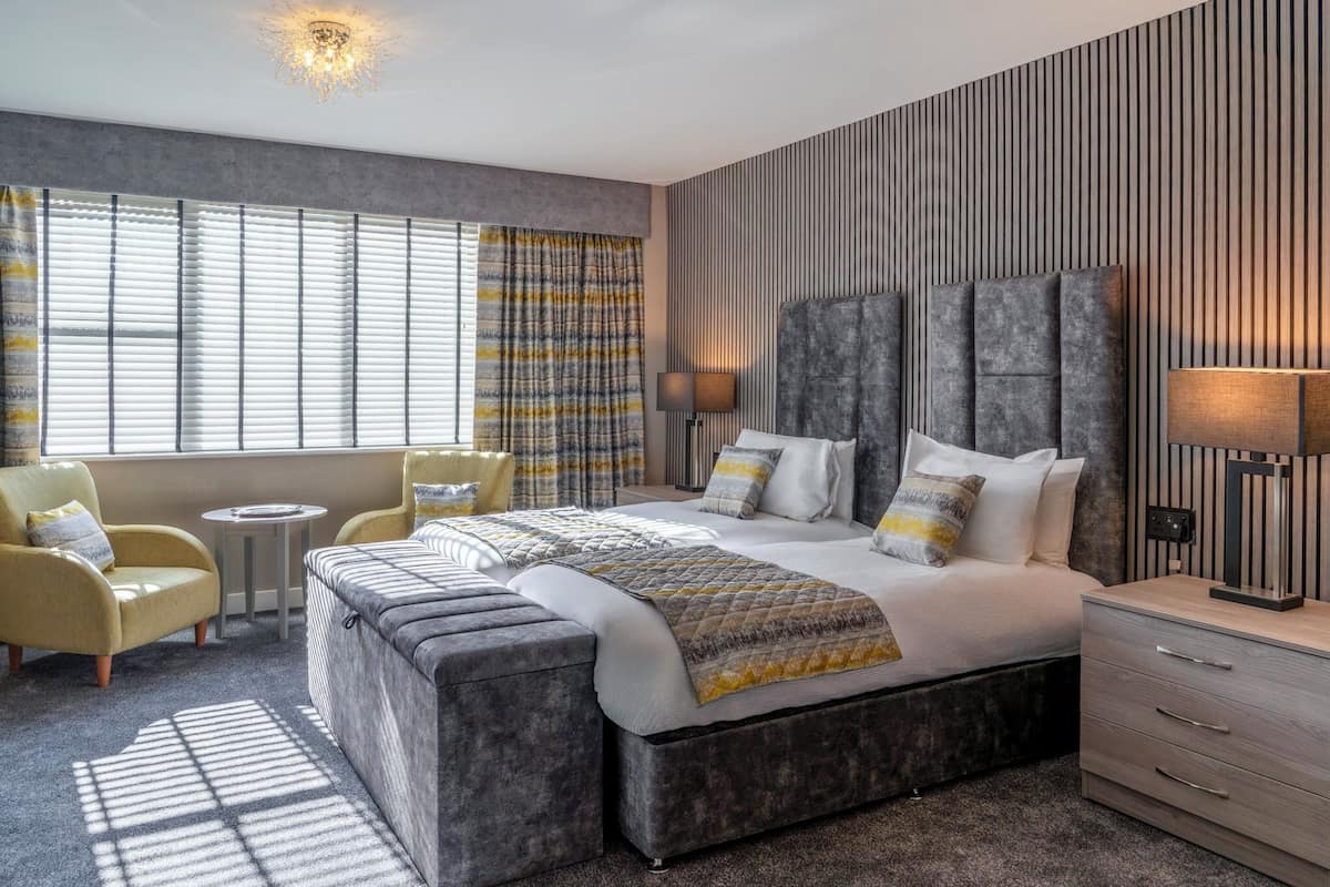 Une chambre d’hôtel moderne dotée d’un grand lit, d’un coin salon avec des fauteuils, d’une petite table et d’une table de chevet avec une lampe.