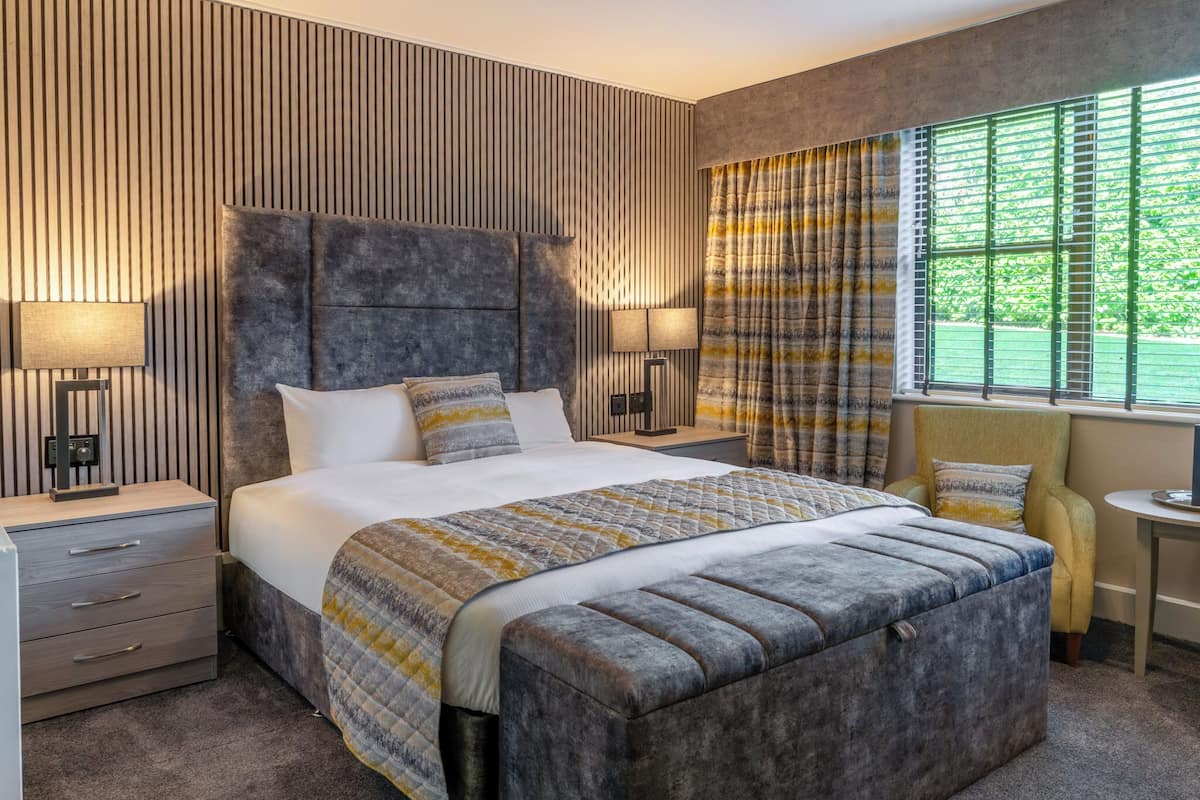 Une chambre d’hôtel moderne avec un grand lit, deux tables de chevet, un petit fauteuil et une fenêtre avec des rideaux.