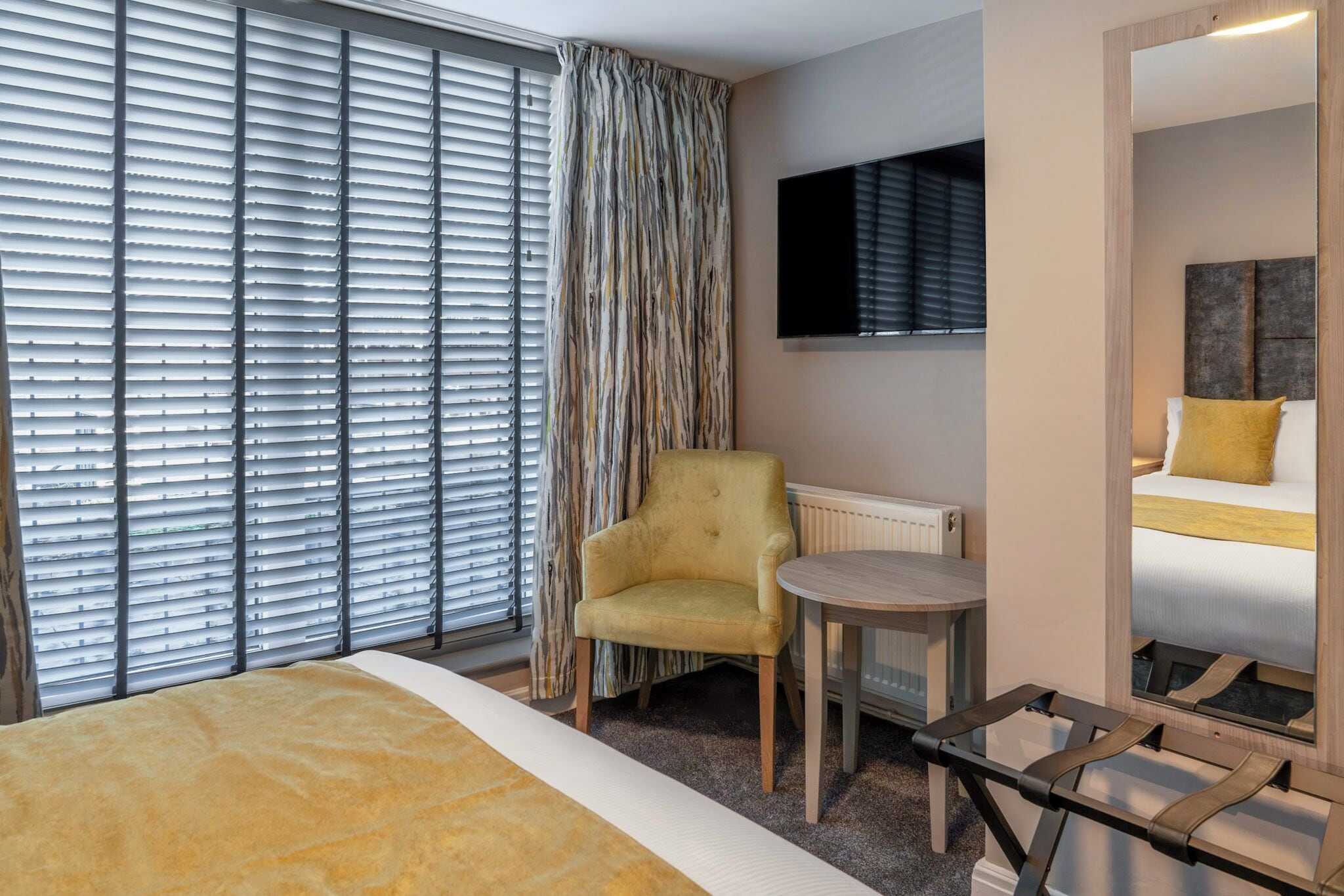 A hotel room with a bed, a chair, a small table, a TV, and a window with blinds.