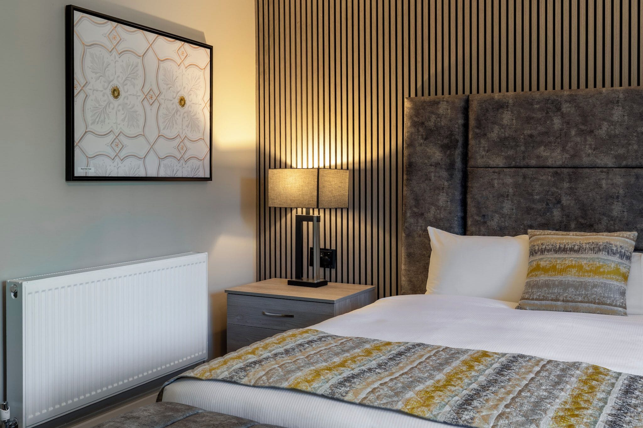 A modern hotel room with a large bed, a bedside lamp, a framed artwork on the wall, and a radiator.