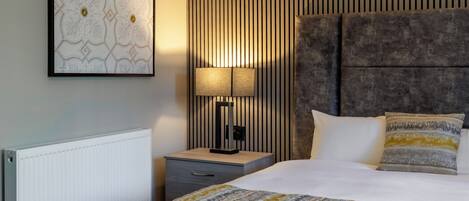 A modern hotel room with a large bed, a bedside lamp, a framed artwork on the wall, and a radiator.