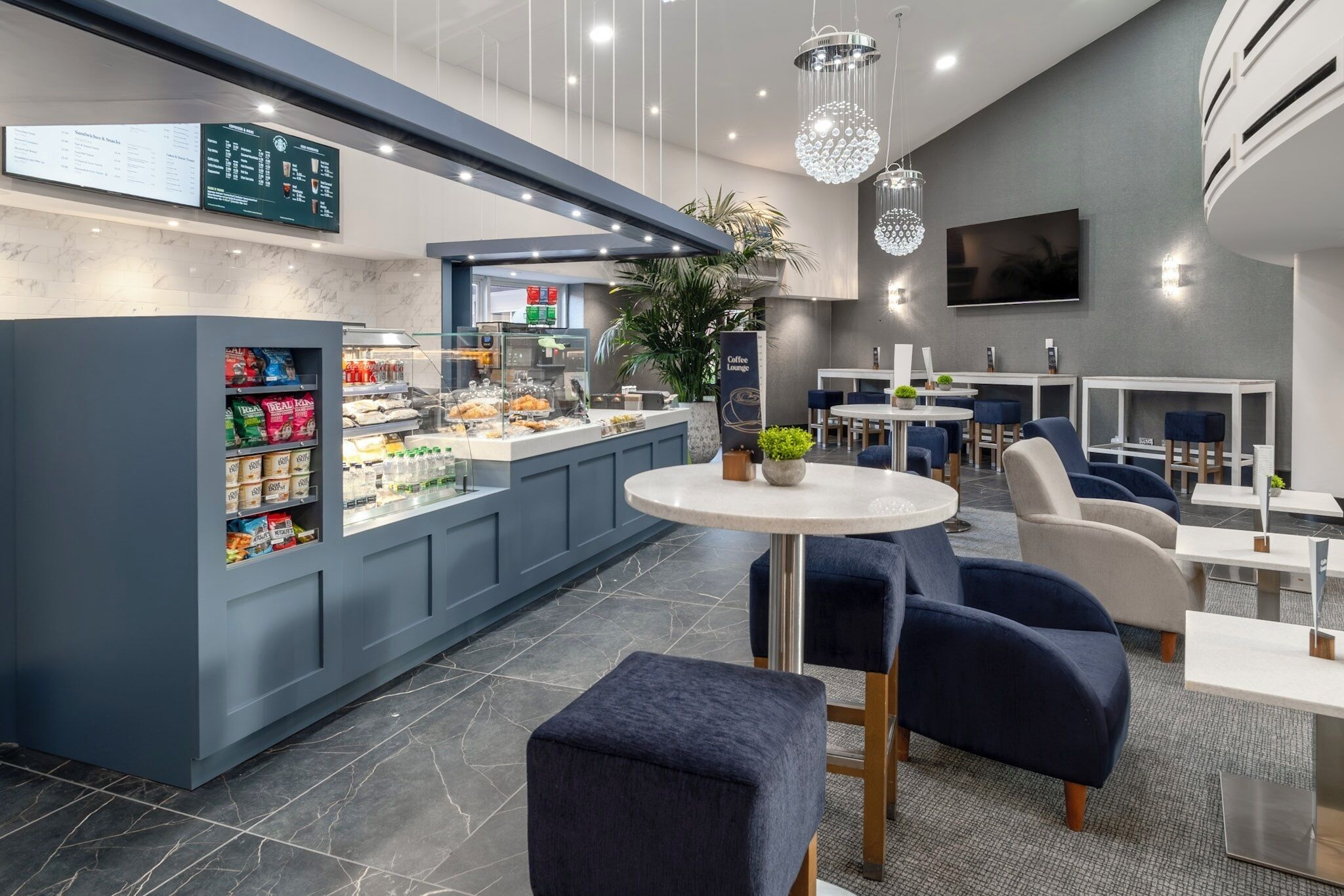 A modern cafe with a counter displaying snacks and drinks, seating area with tables and chairs, and a TV mounted on the wall.