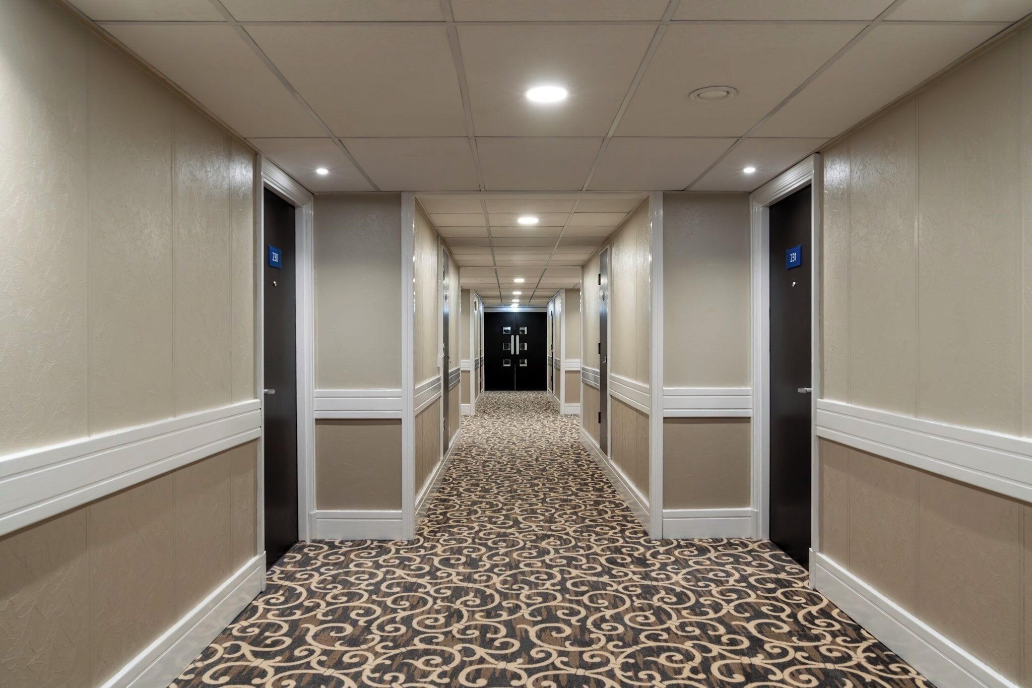 Een hotelgang met een kamergestreept tapijt, crèmekleurige muren en deuren voorzien van kamerNummers.
