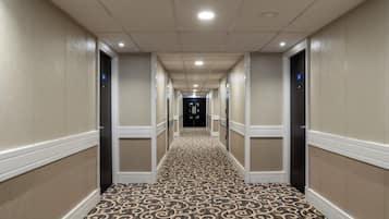 Een hotelgang met een kamergestreept tapijt, crèmekleurige muren en deuren voorzien van kamerNummers.