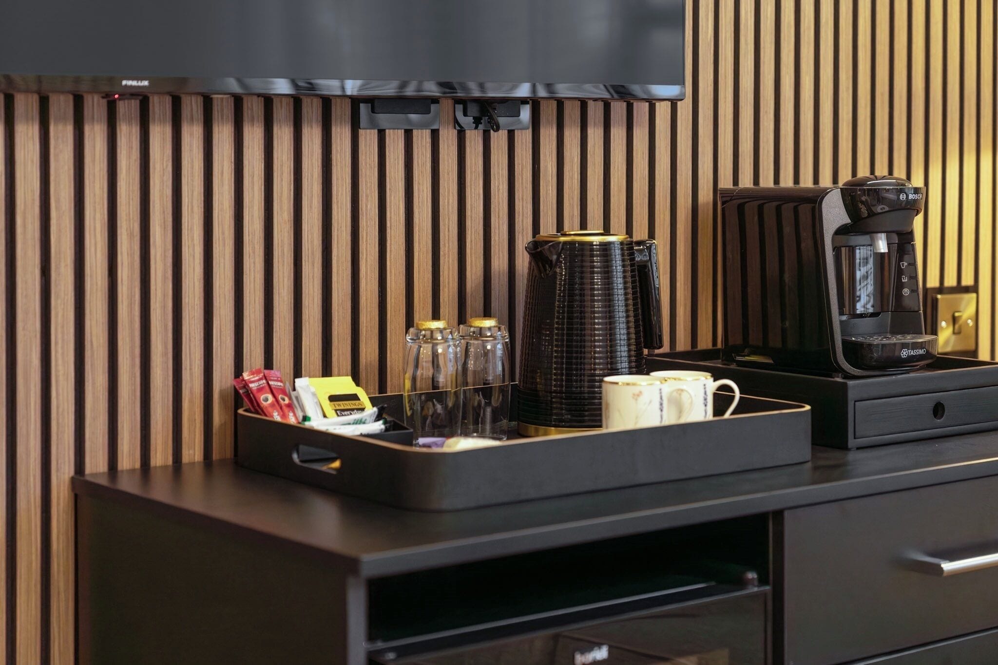 A hotel room with a wooden panel wall, a black coffee maker, a coffee pot, a tray with condiments, and a small table.