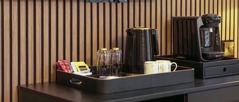 Habitación de hotel con pared de paneles de madera, una cafetera negra, una jarra de café, una bandeja con condimentos y una pequeña mesa.