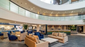 A spacious lobby with a high ceiling, a central seating area with sofas and chairs, a reception desk, and a glass railing overlooking the lower level.