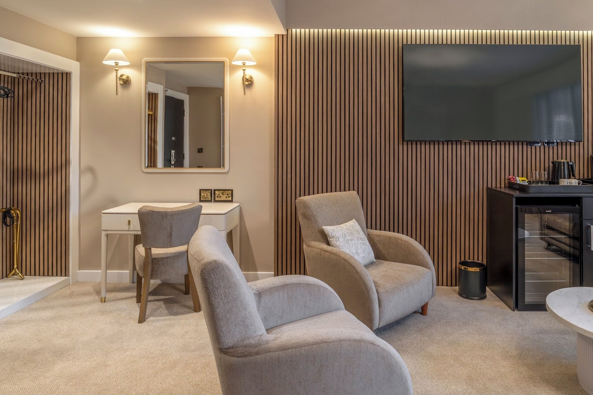 A modern hotel room with a flat-screen TV, a desk with two chairs, a sofa, and a minibar.