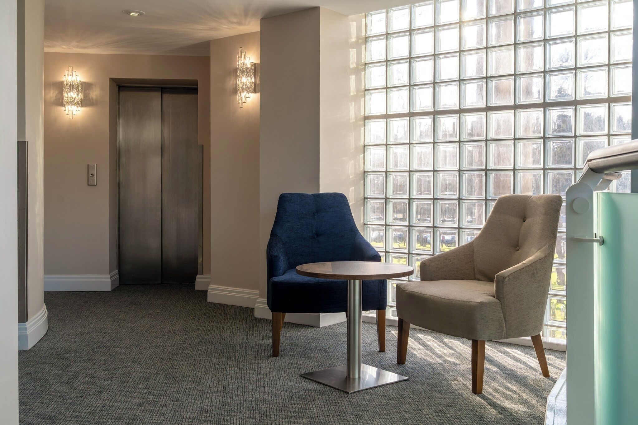 A modern hotel lobby with a large window, two chairs, a round table, and a door leading to an elevator.
