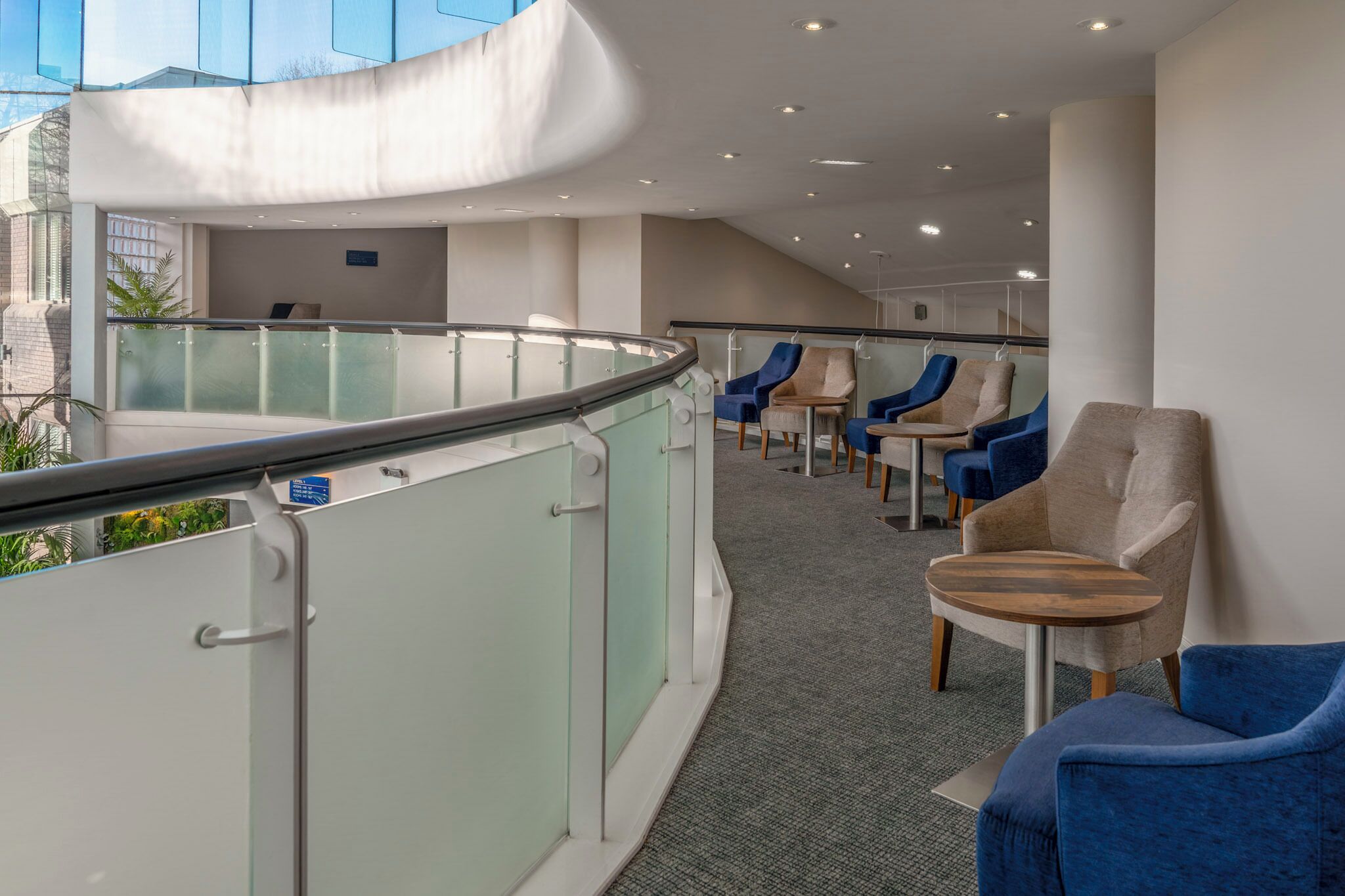 A modern hotel lobby with a curved glass railing, seating area with blue and beige chairs, and a carpeted floor.